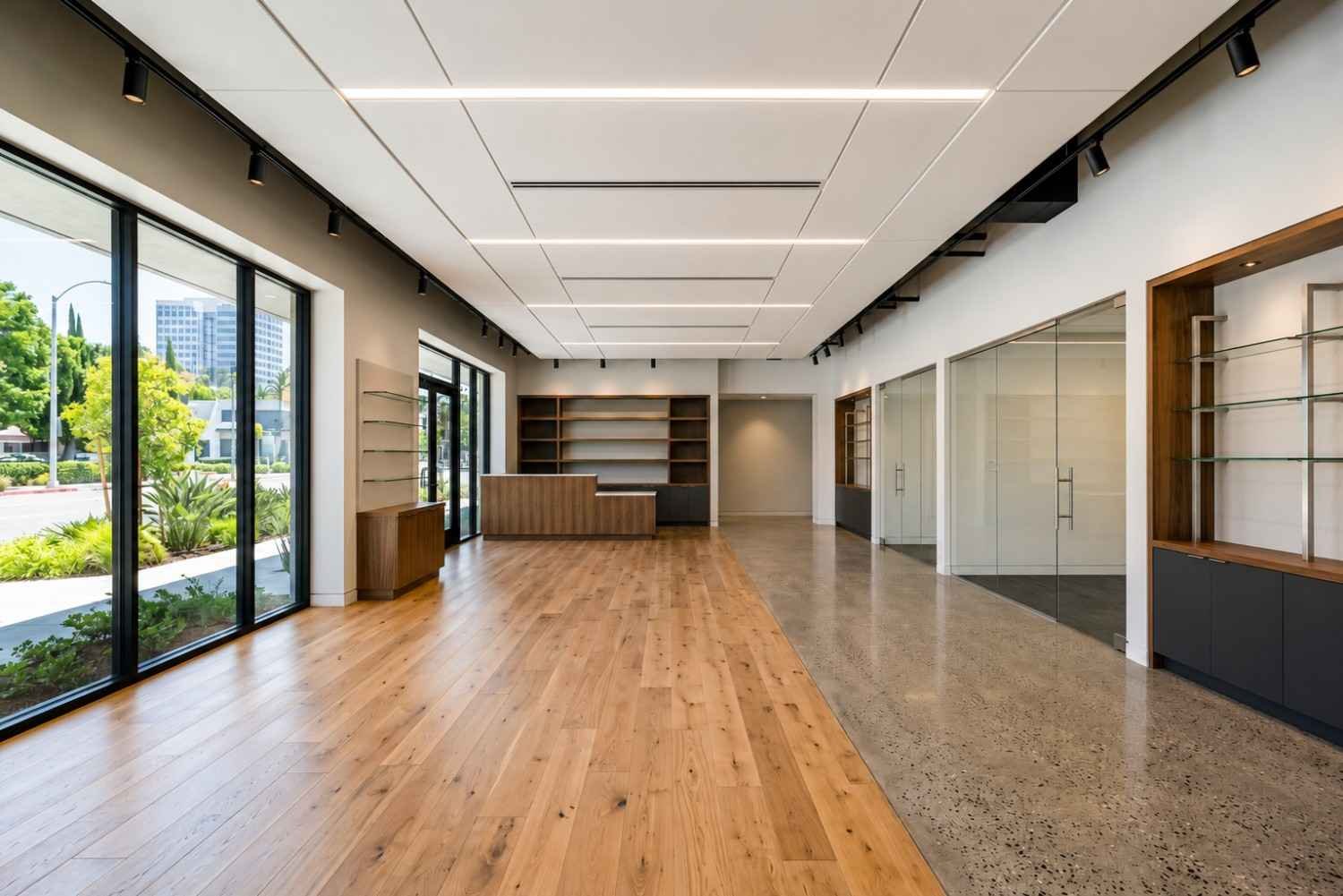 Modern commercial office interior featuring wood flooring, polished concrete, floor-to-ceiling windows, glass offices, custom shelving, recessed LED ceiling lighting, and a minimalist reception area.