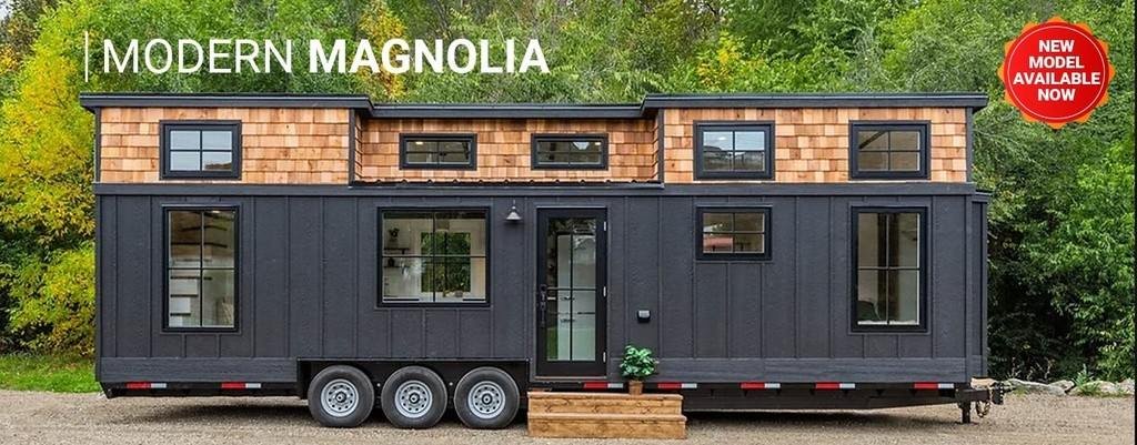 Modern Magnolia tiny home with dark exterior siding, cedar accents, large windows, and a triple-axle trailer surrounded by trees.