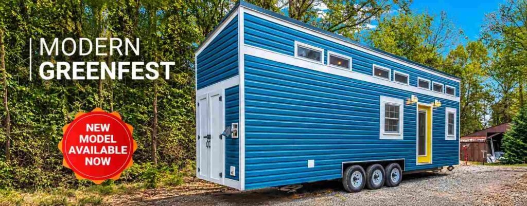 Modern blue tiny home with white trim and triple-axle trailer parked in a wooded outdoor setting.