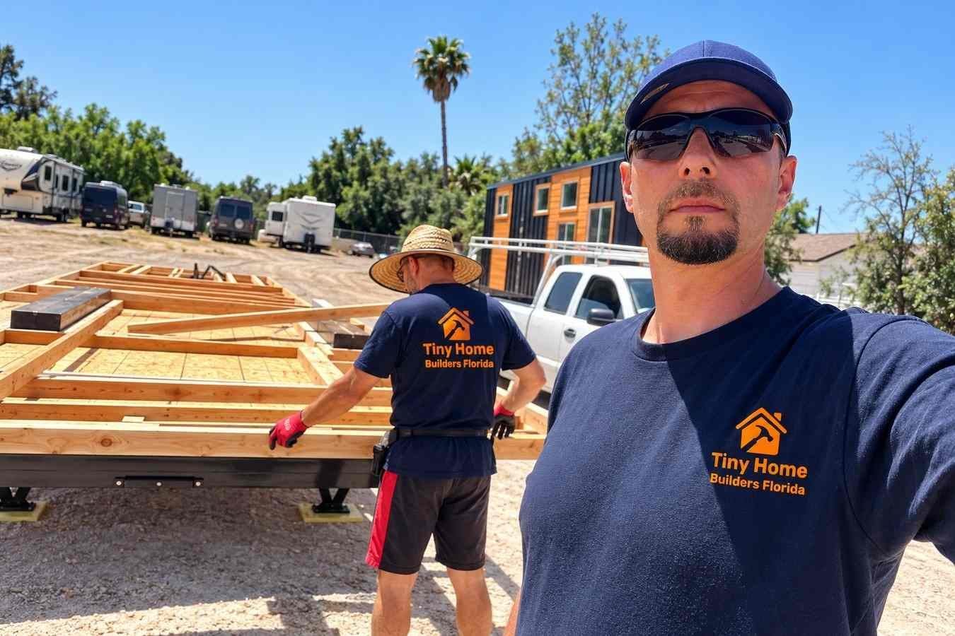 Two construction workers building a tiny home on a raised trailer foundation at an outdoor construction site, with timber floor framing, a white pickup truck, and tiny homes in the background under a clear blue sky.